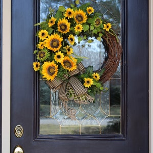 May include: A vibrant sunflower wreath with a brown grapevine base, adorned with lush yellow sunflowers, green leaves, and a checkered ribbon. The wreath hangs on a black door with a glass panel.