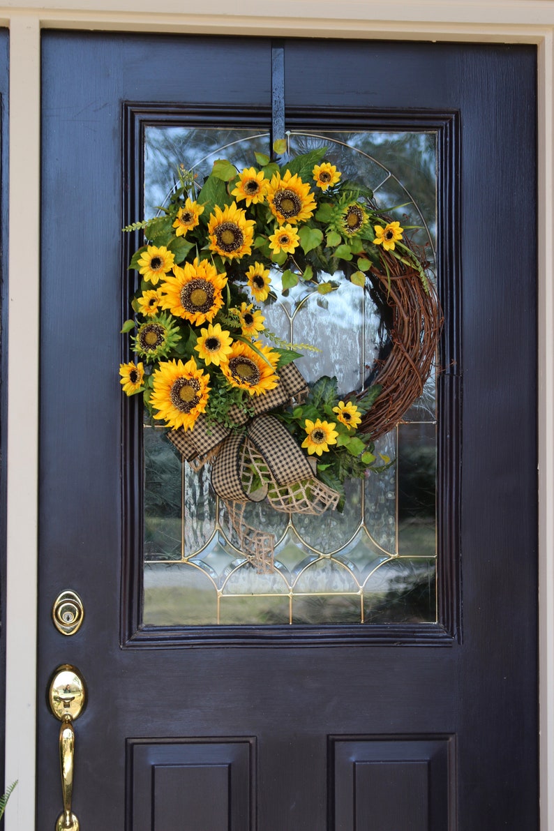 May include: A vibrant sunflower wreath with a brown grapevine base, adorned with lush yellow sunflowers, green leaves, and a checkered ribbon. The wreath hangs on a black door with a glass panel.