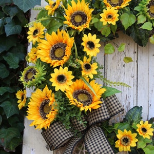 May include: A vibrant sunflower wreath with large yellow blooms and dark centers. The wreath features a black and tan checkered bow and is accented with green leaves. The wreath is displayed on a white, weathered wooden door.