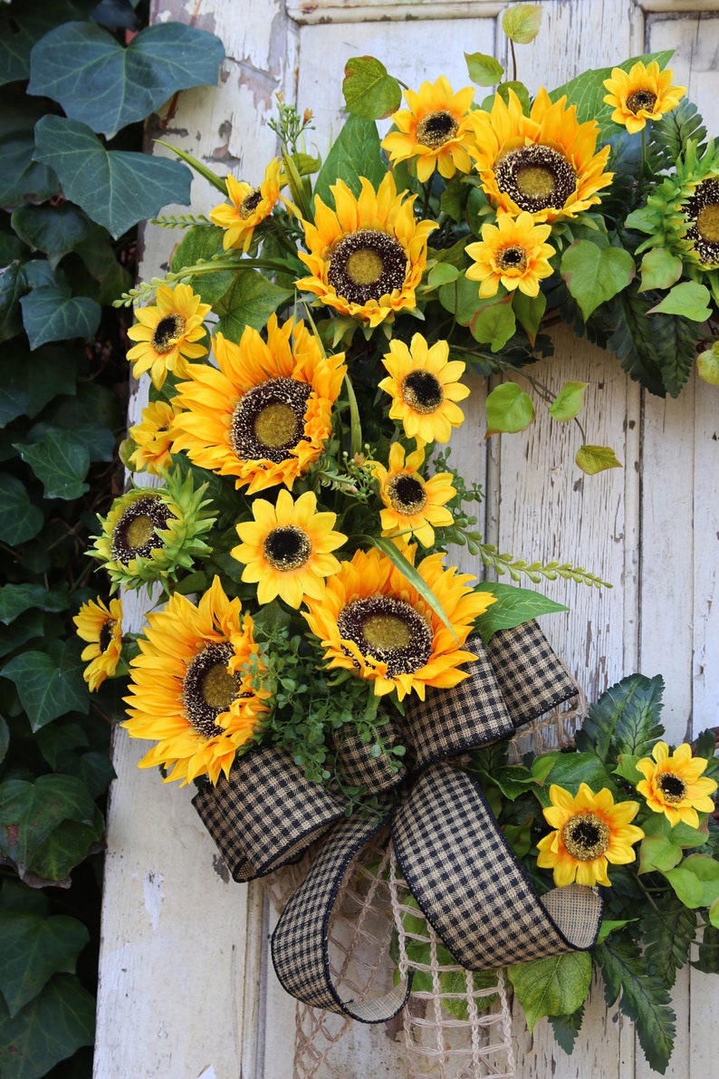 May include: A vibrant sunflower wreath with large yellow blooms and dark centers. The wreath features a black and tan checkered bow and is accented with green leaves. The wreath is displayed on a white, weathered wooden door.