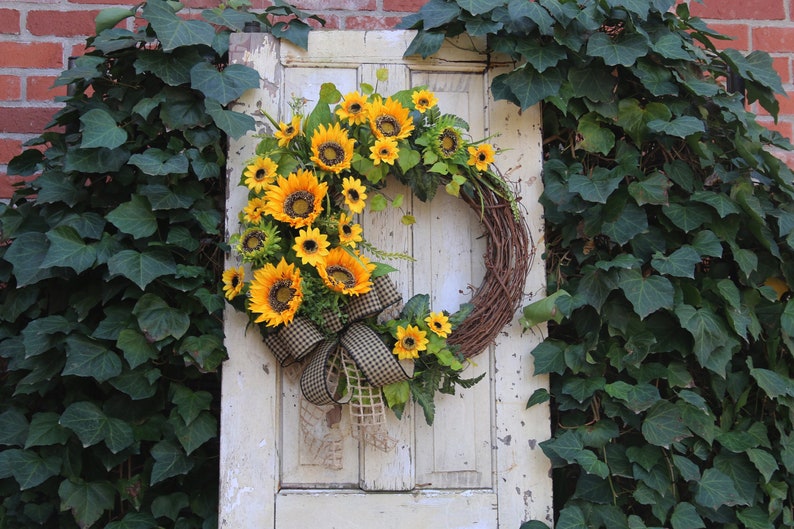 May include: A rustic grapevine wreath adorned with vibrant yellow sunflowers, green leaves, and a checkered ribbon. The wreath is displayed on a distressed white door, surrounded by lush green ivy, creating a welcoming, natural aesthetic.