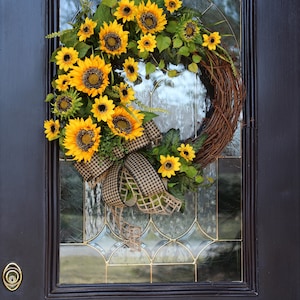May include: A vibrant sunflower wreath with a brown grapevine base, adorned with bright yellow sunflowers, green leaves, and a checkered ribbon bow. The wreath hangs on a dark brown door with glass panels.