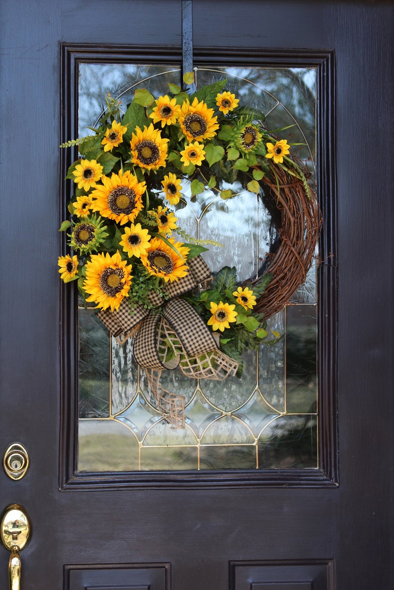 May include: A vibrant sunflower wreath with a brown grapevine base, adorned with bright yellow sunflowers, green leaves, and a checkered ribbon bow. The wreath hangs on a dark brown door with glass panels.