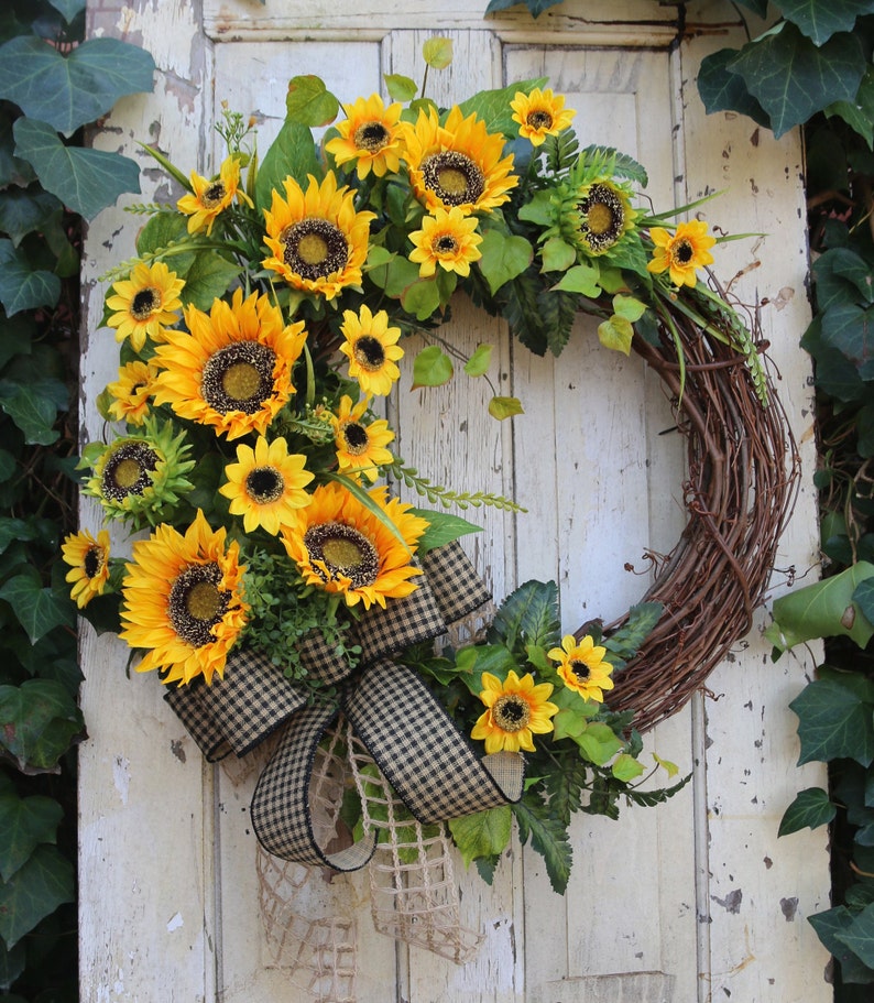 May include: A vibrant sunflower wreath with a rustic, natural grapevine base. The wreath features large, bright yellow sunflowers, green foliage, and a black and tan checkered bow. A decorative accent for doors or walls.