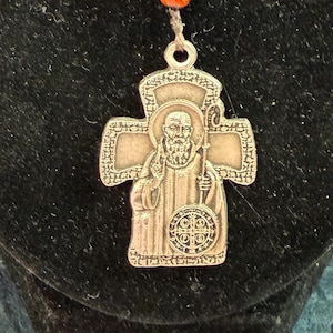 May include: A religious pendant featuring a silver-colored cross with an image of a saint, connected to a series of dark gray, flower-shaped beads and small orange beads. The pendant is attached to a silver-colored clasp.