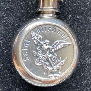 Op de afbeelding: Een zilverkleurige flacon met een scharnierende bovenkant en een sluiting. De flacon heeft een reliëfbeeld van Sint-Michiel met de woorden "Saint Michael" en "Michael" rond het beeld. De flacon is ontworpen om aan een sleutelhanger te worden bevestigd.