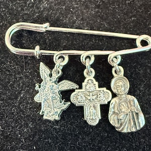May include: Silver safety pin with three religious charms: an angel, a cross, and a saint. The charms feature religious iconography. The pin is a classic design, suitable for various uses.