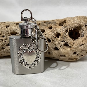 Op de afbeelding: Een kleine, rechthoekige, zilverkleurige flacon met een decoratief hartvormig embleem. Het heeft een schroefdop en een metalen sleutelhangerclip. De flacon wordt getoond tegen een lichtgekleurde, getextureerde houten achtergrond.