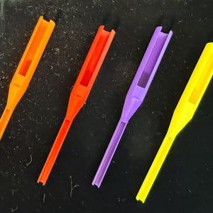 May include: Four plastic tools in vibrant colors: orange, red, purple, and yellow. Each tool has a long, thin handle and a rectangular opening at the top. The tools are arranged in a row against a dark background.