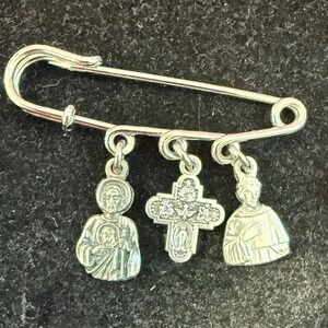 May include: A silver-coloured safety pin with three dangling charms. The charms include two figures and a cross. The figures are detailed with etched designs. The safety pin is a simple, functional design.