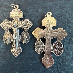 May include: Two ornate silver-toned religious crosses. One features a crucifix with the inscription "JESUS NAZARENUS REX JUDÆORUM." The other cross has the text "BEHOLD THIS HEART" and "FORGIVE THEM."