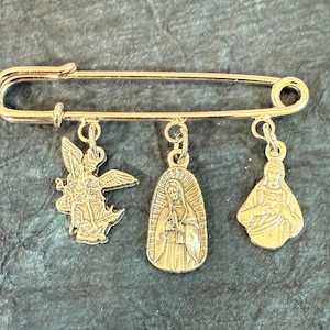 May include: Gold-coloured safety pin with three hanging charms. The charms show an angel, the Virgin Mary, and Jesus. This religious accessory is suitable for attaching to clothing or accessories.