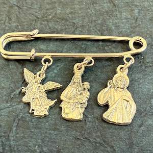 May include: A silver-coloured safety pin with three dangling religious charms. The charms depict an angel, the Virgin Mary with child, and Jesus. The pin is made of a shiny metal against a dark green background.