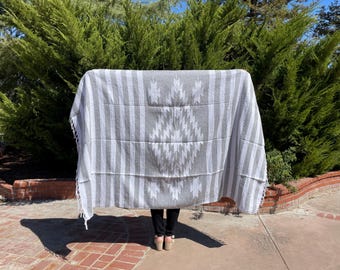 May include: A grey and white woven blanket with a geometric design. The blanket features vertical white stripes and a central diamond pattern. The edges have a fringe detail. The blanket is held up in front of a person.