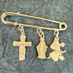 May include: A gold-coloured safety pin with three hanging charms: a crucifix, a depiction of the Virgin Mary, and a heart pierced by arrows. This pin is a religious accessory.