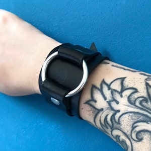 Puede incluir: Pulsera de cuero negro con un anillo de plata. La pulsera se lleva en la muñeca de una persona con un tatuaje de una rosa en blanco y negro.