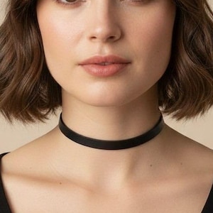 May include: A simple black choker necklace worn around the neck. The choker is a thin, smooth, solid black band that sits flush against the skin. It is paired with a plain black top featuring a wide, rounded scoop neckline.