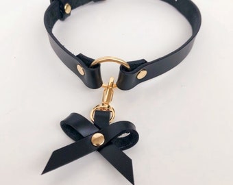 May include: A black leather choker featuring a gold-toned metal O-ring at the center. A detachable black leather bow charm with a gold-toned rivet is attached to the O-ring via a gold-toned lobster clasp. The choker has an adjustable buckle.