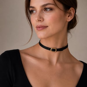 Pode incluir: Um colar gargantilha preto com uma fivela dourada. A gargantilha &eacute; feita de um material fino e liso e &eacute; presa com uma fivela retangular. O colar &eacute; usado em volta do pesco&ccedil;o.