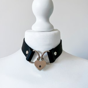 Könnte beinhalten: Ein schwarzes Lederhalsband mit einem silbernen herzförmigen Vorhängeschloss und zwei silbernen Ringen.