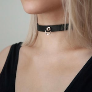 Puede incluir: Un collar gargantilla de cuero negro con un anillo plateado y un acento de bola plateada.