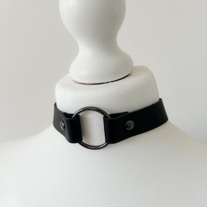 Op de afbeelding: Een zwarte leren choker met een zilveren metalen ring. De choker is op een wit mannequin.
