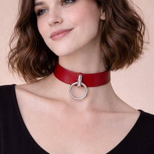 Puede incluir: Un collar gargantilla de cuero rojo con un gran colgante de anillo plateado. El collar se lleva alrededor del cuello. El fondo es de color neutro. Un accesorio de moda llamativo.