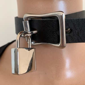 Puede incluir: Collar gargantilla de cuero negro con hebilla cuadrada plateada y colgante de candado a juego. La gargantilla se asegura alrededor del cuello con un cierre de hebilla. El candado está sujeto a la gargantilla con un anillo de metal.