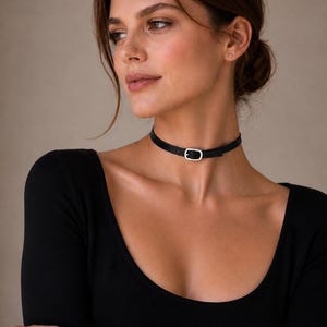 Pode incluir: Um colar gargantilha preto com uma fivela prateada. A gargantilha &eacute; feita de um material fino e preto e &eacute; usada ao redor do pesco&ccedil;o. A fivela tem formato oval e prende o colar. A pessoa est&aacute; usando um top preto de manga comprida.