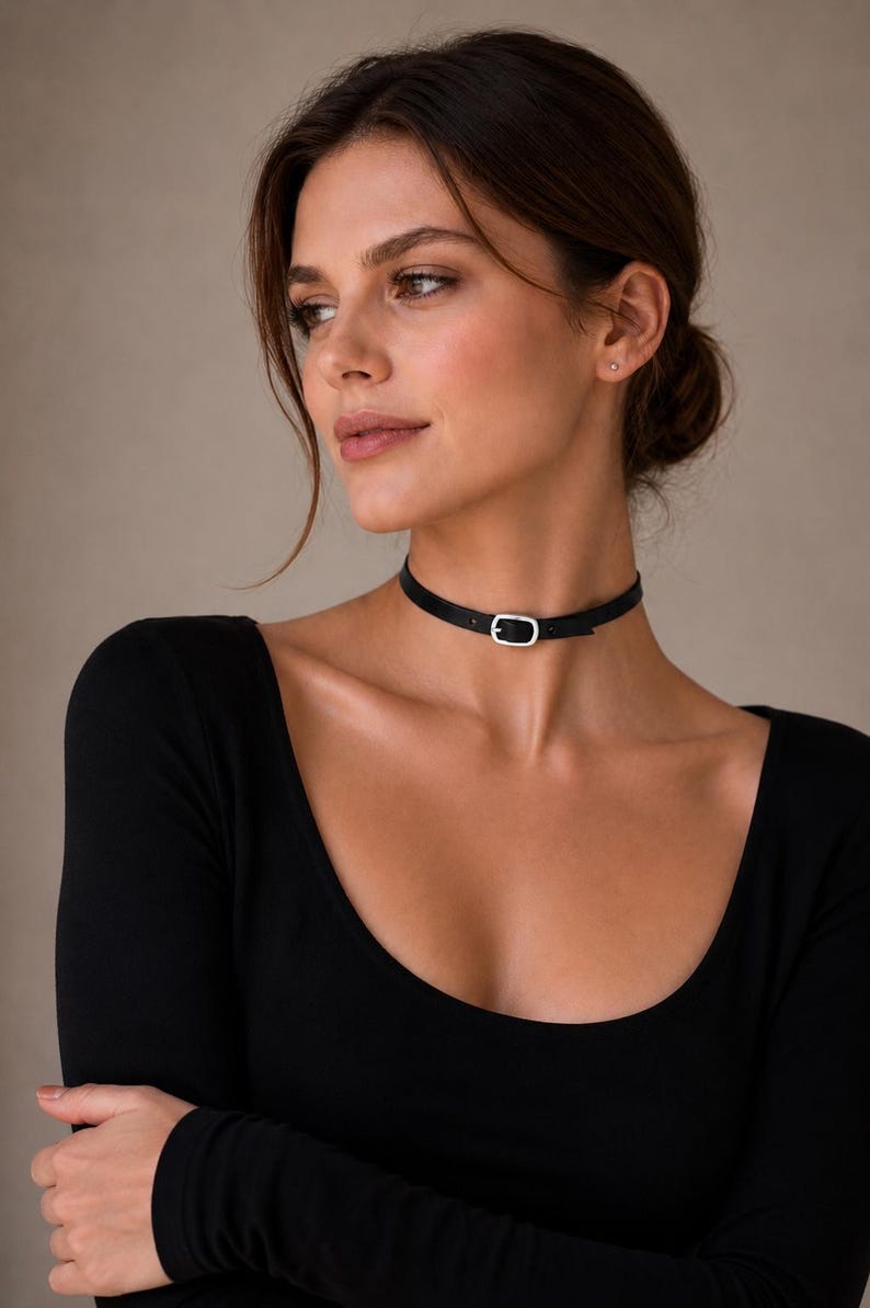 Pode incluir: Um colar gargantilha preto com uma fivela prateada. A gargantilha &eacute; feita de um material fino e preto e &eacute; usada ao redor do pesco&ccedil;o. A fivela tem formato oval e prende o colar. A pessoa est&aacute; usando um top preto de manga comprida.