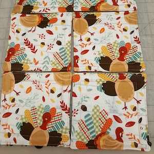 May include: Six fabric coasters with a Thanksgiving theme. Each coaster features a brown and orange turkey with a white background and a pattern of leaves, acorns, and plaid.
