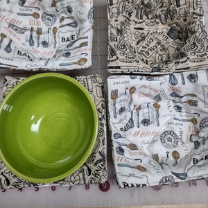 May include: Four white fabric bowl cozies with a black and brown print of kitchen utensils and food-related words. The cozies are folded and arranged on a white grid surface. A green ceramic bowl is sitting on one of the cozies.