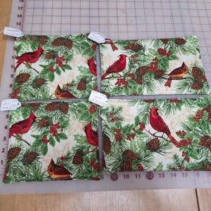May include: Four rectangular fabric coasters featuring a cream background with a pattern of red cardinals, green pine branches, brown pinecones, and red berries. Each coaster has a small white paper tag attached to the corner.