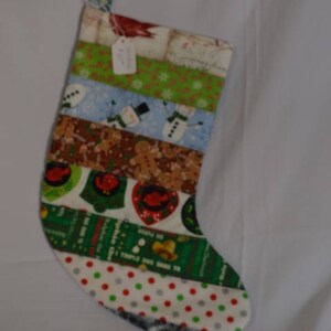 May include: A Christmas stocking made of patchwork fabric with a variety of holiday themes, including gingerbread men, snowmen, and bells. The stocking is green, red, white, and brown.