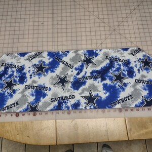 May include: A white table runner with a blue and gray tie-dye pattern. The runner features multiple blue stars and the word "COWBOYS" repeated throughout.