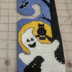 May include: A blue and white cross-stitch door hanger featuring a ghost holding a black bucket, a black cat perched on a branch, and a yellow moon.