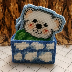 May include: A blue and white crocheted box with a white bear face on top. The bear has a green shirt and red cheeks.