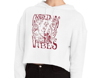Peut inclure: Sweat-shirt blanc à capuche court à manches longues avec un imprimé graphique rouge qui dit "Christmas Vibes" avec un motif circulaire représentant un sapin de Noël et d'autres symboles de Noël.