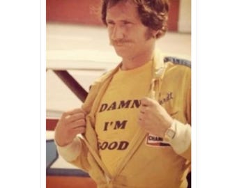Peut inclure: Un homme en combinaison jaune avec un t-shirt jaune qui dit "DAMN I'M GOOD" ouvre son t-shirt pour montrer le texte. Il porte une montre au poignet gauche.