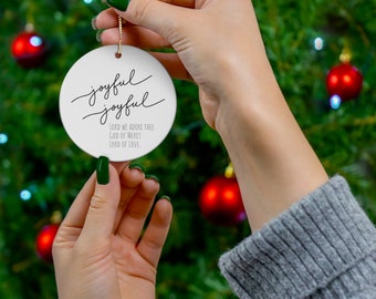 Peut inclure: Ornement de Noël en céramique blanche avec les mots "joyful joyful" écrits en cursive noire. L'ornement comporte également le texte "Lord we adore thee, God of mercy, Lord of love" écrit en noir.