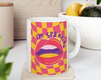 Peut inclure: Une tasse en céramique blanche avec un motif à carreaux rose et jaune. La tasse a une grande bouche rouge avec des lèvres violettes et le texte "I'm Speaking" en lettres violettes.