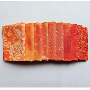 May include: A collection of folded orange fabric squares with various patterns, including floral, geometric, and abstract designs. The fabrics range in shades from light yellow-orange to deep red-orange.