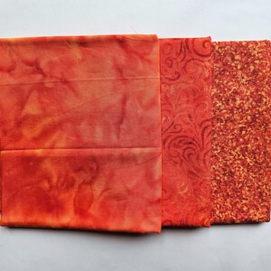 May include: Three folded pieces of fabric in shades of orange and red. The first piece has a mottled, tie-dye appearance. The second piece features a dark red swirling pattern. The third piece has a dense, textured, speckled orange and red print.