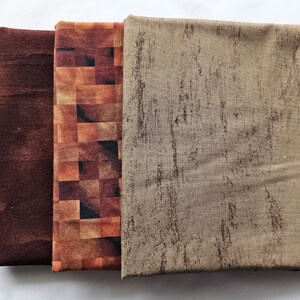 May include: Three folded fabric pieces in shades of brown. The first is a dark, mottled brown. The second has a brick-like pattern in shades of brown and orange. The third is a light brown with a textured, wood-grain appearance.