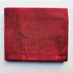 Op de afbeelding: Een klein, vierkant rood stoffen item met een getextureerd oppervlak. De kleur is een diep, rijk rood met donkerdere gebieden, wat wijst op een mogelijk kleureffect of patroon. Het item lijkt gemaakt van geweven materiaal.