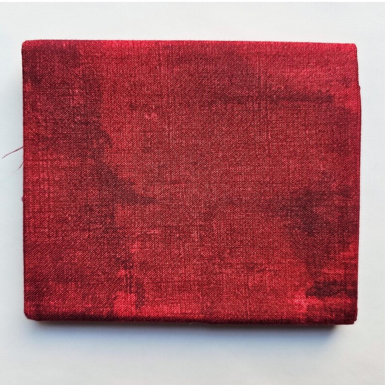 Op de afbeelding: Een klein, vierkant rood stoffen item met een getextureerd oppervlak. De kleur is een diep, rijk rood met donkerdere gebieden, wat wijst op een mogelijk kleureffect of patroon. Het item lijkt gemaakt van geweven materiaal.