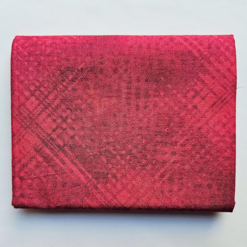 Op de afbeelding: Een rechthoekig, gevouwen stoffen item in een dieprode kleur, met een subtiel, donker kruispatroon. De textuur lijkt enigszins ruw, met een matte afwerking. Het item is waarschijnlijk een portemonnee of een kleine etui.