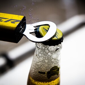 May include: A yellow bottle opener with the word "SUPER" printed on it is being used to open a bottle of beer. The bottle is clear glass and has a yellow label. There are bubbles forming in the beer as the cap is removed.