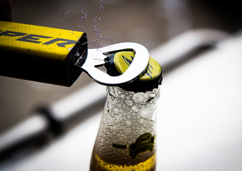 May include: A yellow bottle opener with the word "SUPER" printed on it is being used to open a bottle of beer. The bottle is clear glass and has a yellow label. There are bubbles forming in the beer as the cap is removed.