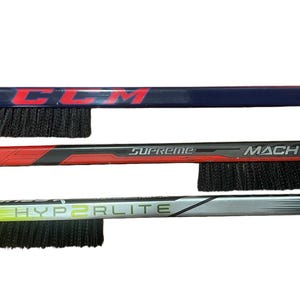 May include: Three hockey stick ice scrapers with black bristles. The top scraper is blue with the word "CCM" printed on it. The middle scraper is red with the words "Supreme" and "MACH" printed on it. The bottom scraper is gray with the words "HYPERLITE" printed on it.
